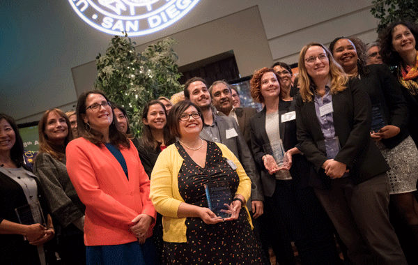 shot from a low, left angle of a group of people posing for the shot. The group is all women of different ages, ethnicities, and heights. There is also one white, younger man and the Chancellor. They are the winners of the 2017 UC San Diego Equal Opportunity/Affirmative Action Awards